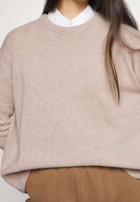 Jasno beżowy sweter z okrągłym dekoltem, miękka faktura i luźny krój, zestawiony z białą koszulą z kołnierzykiem pod spodem. Widoczne detale wykończenia.
