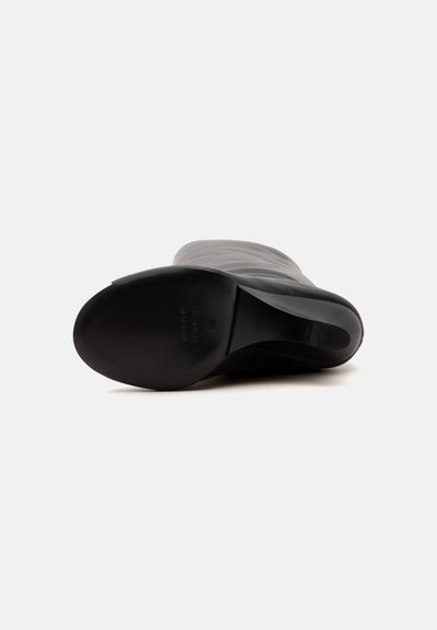 Botte haute noire en cuir, couchée sur le côté, montrant une semelle lisse et un petit talon carré sur fond blanc.