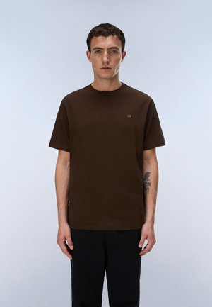 Ung mand med kort mørkt hår iført en brun kortærmet T-shirt og sorte bukser, stående mod en ensfarvet lys baggrund.