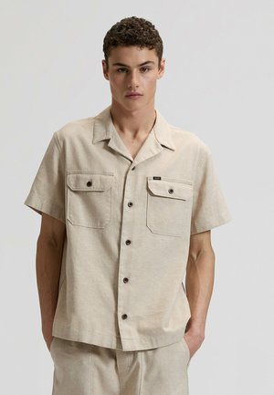 Camicia beige a maniche corte in tessuto leggero con due tasche sul petto, chiusura frontale con bottoni e vestibilità rilassata. I bottoni marroni creano un contrasto.