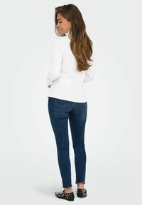 Giacca bianca aderente con maniche lunghe, abbinata a jeans skinny blu scuro. Ballerine nere con pois bianchi, texture liscia e design curato.
