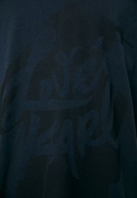 Camiseta de algodón azul oscuro con un diseño gráfico sutil en tono sobre tono que presenta el texto "Love Karl" en una fuente bold cursiva.