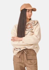 Suéter de punto beige con diseño de flores en lentejuelas, combinado con pantalones cargo marrones. El modelo lleva una gorra color arena, con visera recta.