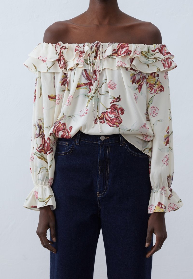 Blouse épaules dénudées crème à motif floral, col à volants et manches longues. Associée à un jean en denim foncé taille haute.