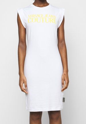 Kvinde iført en ærmeløs hvid kjole med gul "VERSACE JEANS COUTURE" tekst på tværs af brystet og en lille sort logomærkat nær kanten.