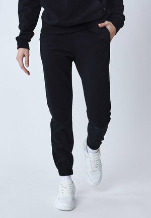 Pantaloni tuta neri con polsini elasticizzati, dotati di una tasca laterale e dettagli del logo in rilievo. Abbinati a scarpe da ginnastica bianche.