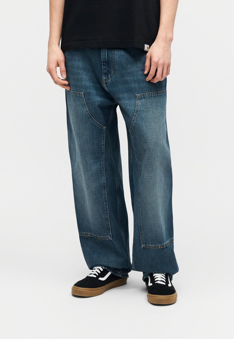 Persona con pantalones sueltos de mezclilla azul con grandes bolsillos delanteros de parche y zapatillas negras con cordones blancos y suelas de goma, de pie sobre un fondo blanco.