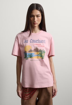 Frau mit langen dunklen Haaren trägt ein pinkfarbenes T-Shirt mit einer Strand-Szene bei Sonnenuntergang mit Bergen, Palmen und französischem Text.