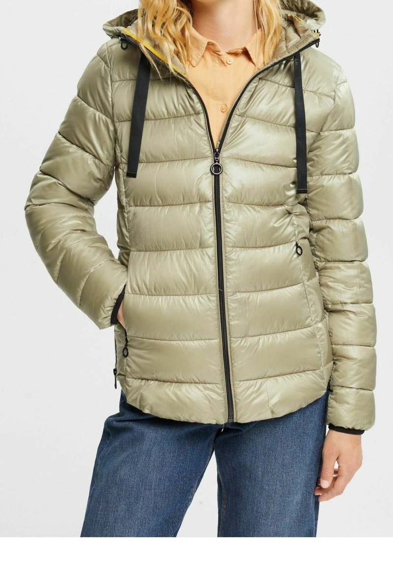 Femme portant une veste matelassée à capuche vert clair avec des cordons noirs sur une chemise beige et un jean bleu, mains dans les poches de la veste.