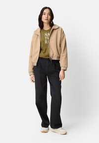 Beige fleece jacket with a zip and ribbed cuffs, green t-shirt with "NATURE" text, black trousers, and white sneakers.
