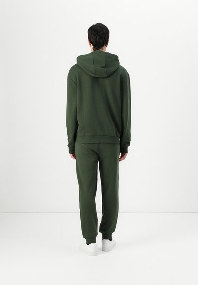 Un sweat à capuche vert foncé et un jogger assorti, fabriqués dans un tissu doux avec une coupe décontractée, comportant une capuche et des poignets élastiques. Des baskets blanches sont portées.