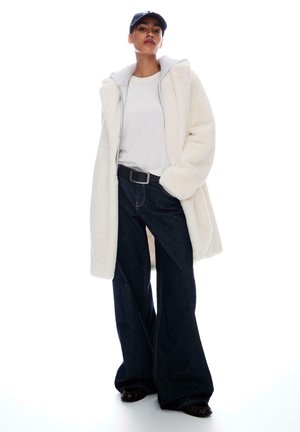 Witte fleecjas over een grijze hoodie, gecombineerd met een wit T-shirt en donkere flared jeans. De outfit bevat een zwarte riem en glanzende schoenen.