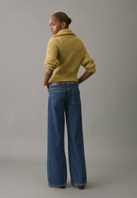 Suéter amarillo mostaza de punto con un cuello alto y mangas enrolladas, combinado con jeans de pierna ancha en azul, visto desde atrás.