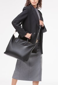 Borsa a tracolla nera in pelle con una texture liscia, due manici corti e una tracolla removibile. Caratterizzata da finiture in metallo dorato e chiusura con zip.
