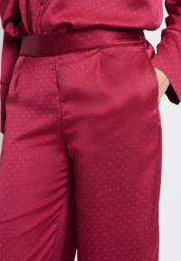 Satijnen bordeauxrode broek met een gladde textuur, voorzien van kleine ruitpatronen en zijzakken. Tailored fit met een hoge taille.