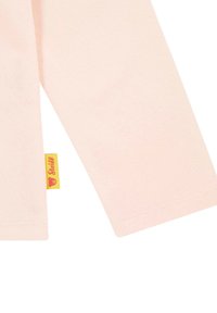 Camisa de manga larga de color rosa hecha de tela suave, con una etiqueta amarilla cosida con un logo de oso cerca de la costura del brazo izquierdo.