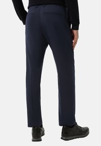 Granatowe spodnie garniturowe wykonane z gładkiej tkaniny, o prostym kroju, z minimalnymi zdobieniami i dwoma tylnymi kieszeniami. Noszone z czarnymi butami.