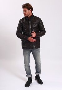 Braune Lederjacke mit hohem Kragen, vorderem Reißverschluss und detaillierten Nähten. Getragen über hellblauen Jeans und schwarzen Knöchelstiefeln.