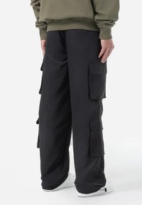 Black cargo pants with large side pockets, loose fit, elastic waistband, and drawstring detail at the ankles, paired with white sneakers.