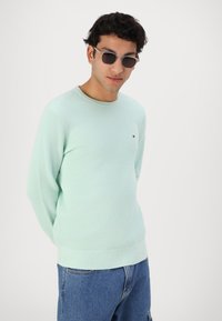 Pull en maille vert clair avec des poignets et un ourlet côtelés, porté avec un jean en denim bleu et accompagné de lunettes de soleil noires.