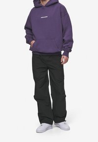 Felpa viola con logo "PEGADOR", tasca a marsupio frontale, abbinata a pantaloni neri larghi e sneakers bianche.