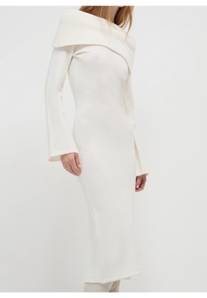 Kobieta w białej dzianinowej sukience z długim rękawem i odkrytymi ramionami, z rozkloszowanymi mankietami, stojąca na jasnym, jednolitym tle.