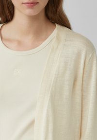 Cardigan beige realizzato in un tessuto leggero e texturizzato, indossato sopra una maglietta crema a coste con un piccolo logo ricamato sul petto.