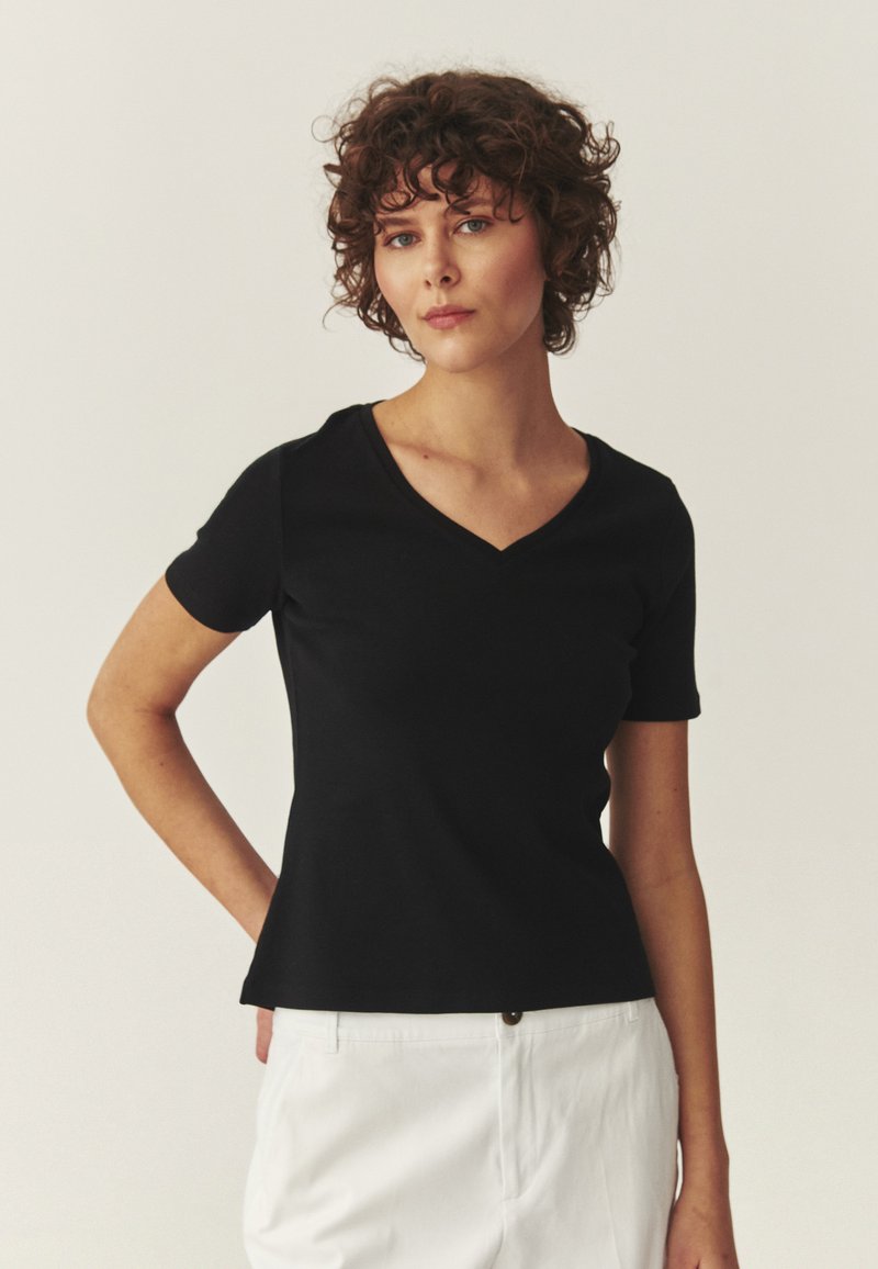 Femme aux cheveux bouclés portant un haut noir à manches courtes et col en V, ainsi qu'un pantalon blanc, debout avec une main derrière le dos.