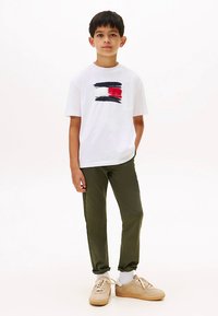 White cotton t-shirt with navy, red, and white brushstroke design; green slim-fit trousers; beige sneakers with white ankle socks.