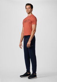 De outfit voor mannen bestaat uit een oranje T-shirt met zwart logo en marineblauwe joggingbroek met elastische tailleband, gecombineerd met zwarte sportschoenen.