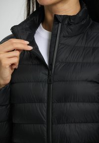 Sorte puffjakke med høj krave og lynlås foran. Har vandrette quiltede sektioner og en glat, skinnende overflade.