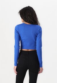 Blue ribbed crop top with long sleeves and a round neckline, paired with high-waisted black pants. Back view shows cropped length and textured fabric.