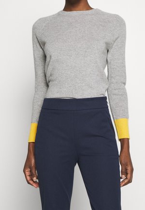 Woman wearing a light gray long-sleeve sweater with yellow cuffs and navy blue high-waisted trousers, standing against a white background.