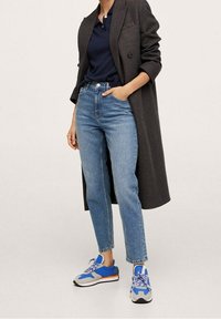 Jeans blu a vita alta, polo blu navy e un lungo cappotto nero con motivo. Sneakers sportive blu, grigie e arancioni con stringhe.