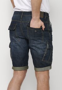 Homme portant un short cargo en denim bleu foncé avec des ourlets roulés et plusieurs poches à l’arrière et sur les côtés, main dans la poche arrière.