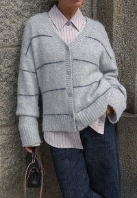 Hellgrauer Cardigan mit blauen Streifen, strukturierter Stoff, Knopfverschluss und übergroßen Ärmeln, kombiniert mit einem gestreiften Hemd und dunklen Jeans.