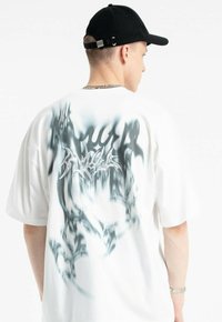 Biała, oversize'owa koszulka z gradientowym czarnym wzorem na plecach. Materiał wydaje się miękki, o gładkiej teksturze.