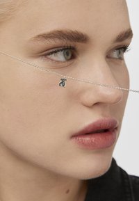 Cadena de plata con un colgante de oso de peluche con acentos negros, reposando cerca de la nariz del modelo. Tono de piel natural y color de labios suave visibles.