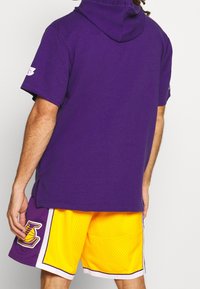 Uomo che indossa un hoodie viola a maniche corte e pantaloni da basket gialli con il logo viola dei Lakers sul lato sinistro, visto da dietro.