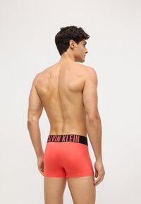 Muške bokserice u svijetlo koraljnoj boji s crnim pojasom na kojem piše "CALVIN KLEIN" u podebljanim crvenim slovima, glatke teksture, prilagođenog kroja.