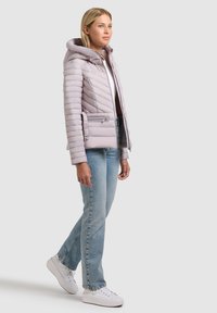 Naine, kes kannab helehallis polsterdatud kapuutsiga jopet, siniseid teksapükse ja valgeid tossusid, seisab ühe jalaga veidi eespool halli tausta ees.