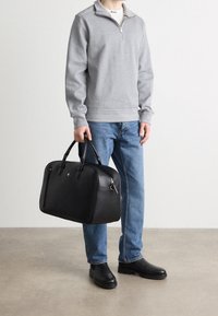 Black leather duffel bag with rounded shape, dual handles, silver hardware, and subtle embossed logo, paired with a gray sweatshirt and blue jeans.