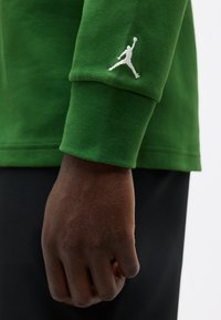 Gros plan d'une main à la peau foncée contre la manche d'un sweat-shirt vert avec un logo en silhouette blanche, tenant un ballon de basket au niveau du poignet.