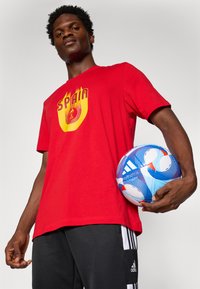 Rød bomulds T-shirt med "Spanien" grafik i gule flammer. Modellen holder en blå og hvid fodbold, iført sorte atletiske shorts med striber.