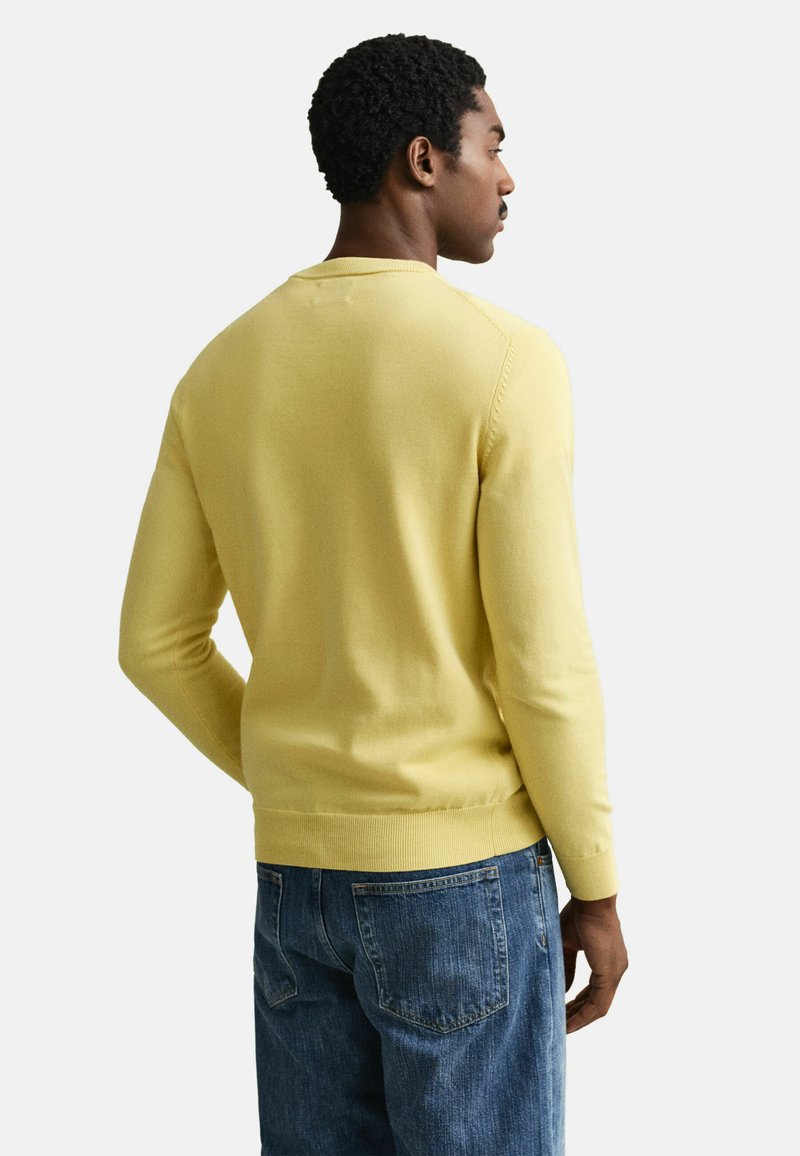 Mann steht mit dem Rücken zur Kamera, trägt einen hellgelben Pullover und blaue Jeans vor einem schlichten weißen Hintergrund.