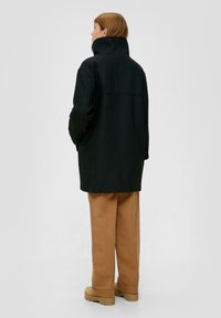 Zwarte wolmix jas met hoge kraag, verlaagde schouders en een relaxte pasvorm, gecombineerd met beige wijde broek en stevige schoenen.