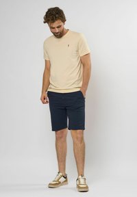 Tricou bej cu mânecă scurtă și un logo mic, asociat cu pantaloni scurți bleumarin. Pantofii sport albi și bej au accente închise la culoare și tălpi texturate.