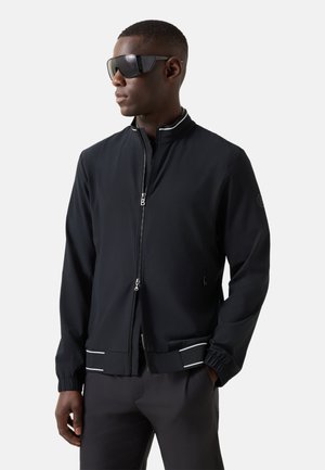 Man die een zwarte jas met rits en witte bies draagt en een donkere zonnebril op heeft, staand tegen een effen lichte achtergrond met handen in de zakken.