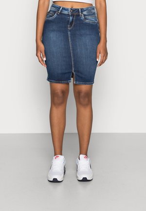 Kvinde iført en knælængde blå denimnederdel med frontslids og hvide Nike Air-sneakers foran en ensfarvet baggrund.