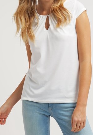 Haut blanc à manches courtes avec une ouverture en forme de goutte d'eau au niveau du col, fabriqué en tissu doux, associé à un jean bleu clair. Texture lisse, design simple.